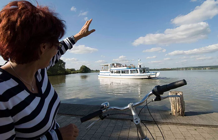 Eine Fahrradfahrerin grüßt die Passagiere der MS Altmühlsee vom Ufer aus