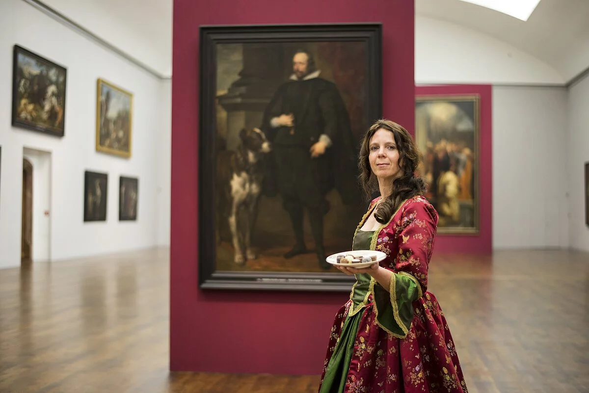 „Kulinarische Reise durch den Barock“ in Neuburg a.d. Donau Eine Frau in einem historischem Kleid trägt einen weißen Teller mit Essen in einer Hand. Im Hintergrund ist ein Porträt von Ottheinrich im Residenzschloss zu sehen.
