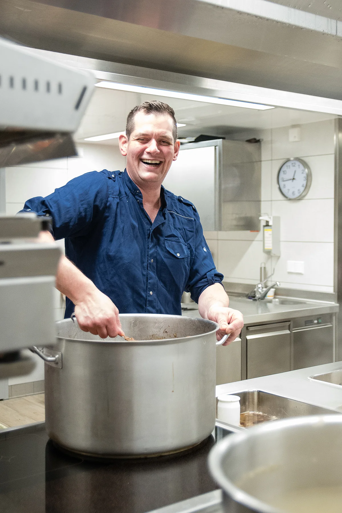 Andreas Blob vom Gasthaus Ziegelhütte Andreas Blob lacht freudestrahlend und rührt in einem riesigen silbernen Kochtopf. Die restliche Küche ist leer, mit weißen Fliesen und silbernen Arbeitsplatten ausgestattet.
