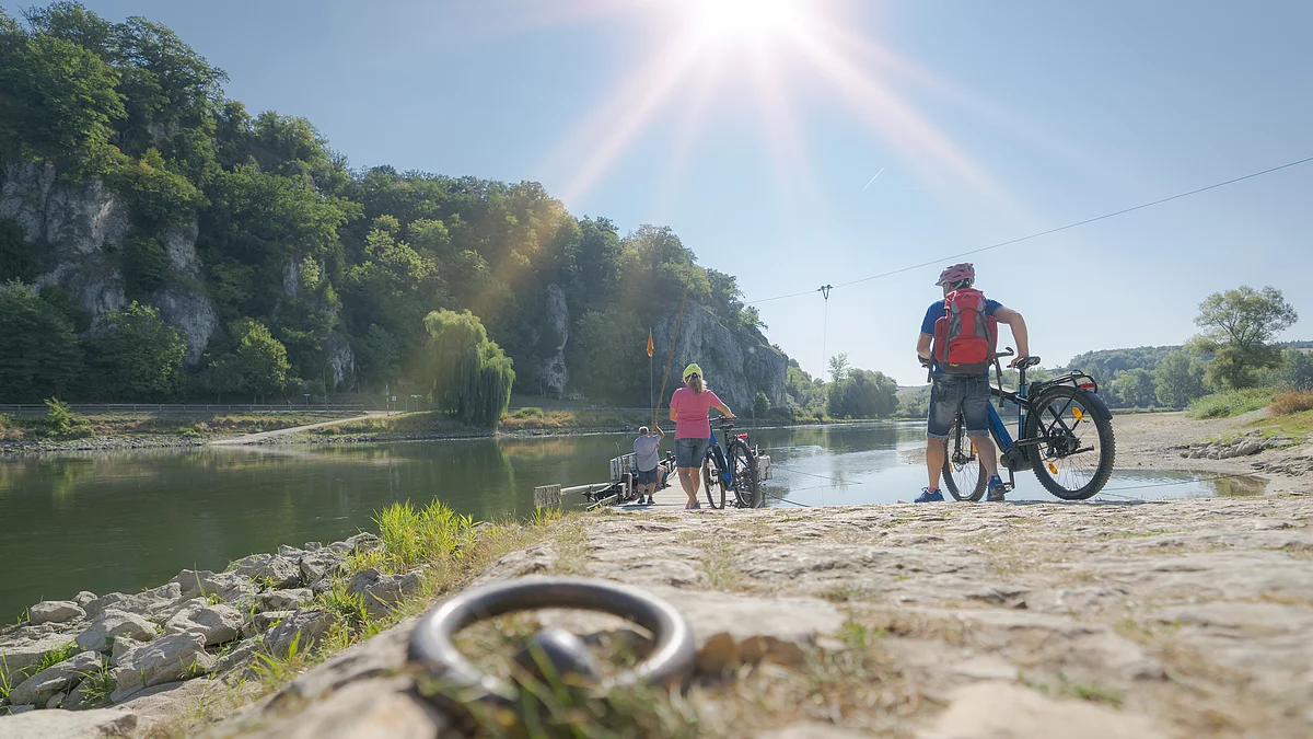 Radler an der Donaufähre bei Weltenburg Personen mit Fahrrädern stehen an einem Flussufer mit bewaldeten Felsen im Hintergrund bei Sonnenschein.