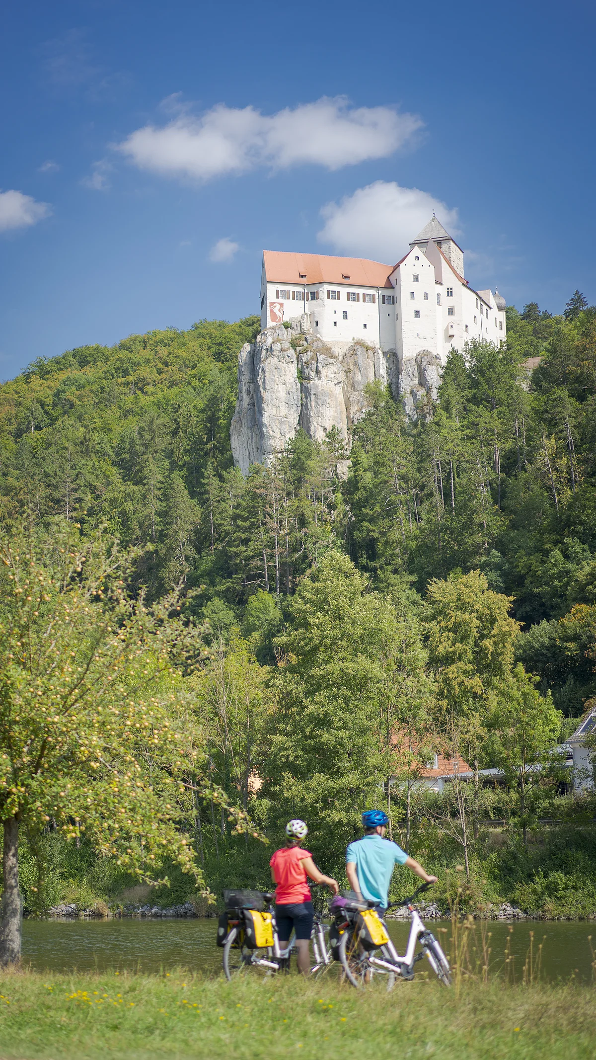 Radler unterwegs auf dem Altmühltal-Radweg bei Burg Prunn Zwei Radfahrer stehen an einem Fluss, im Hintergrund ein weißes Schloss auf einem Felsen und bewaldete Hügel.
