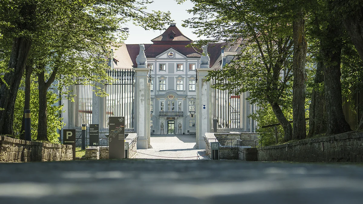 Schloss Hirschberg Eingangstor zu einem historischen Gebäude mit Bäumen und Schildern am Wegesrand bei Tageslicht.