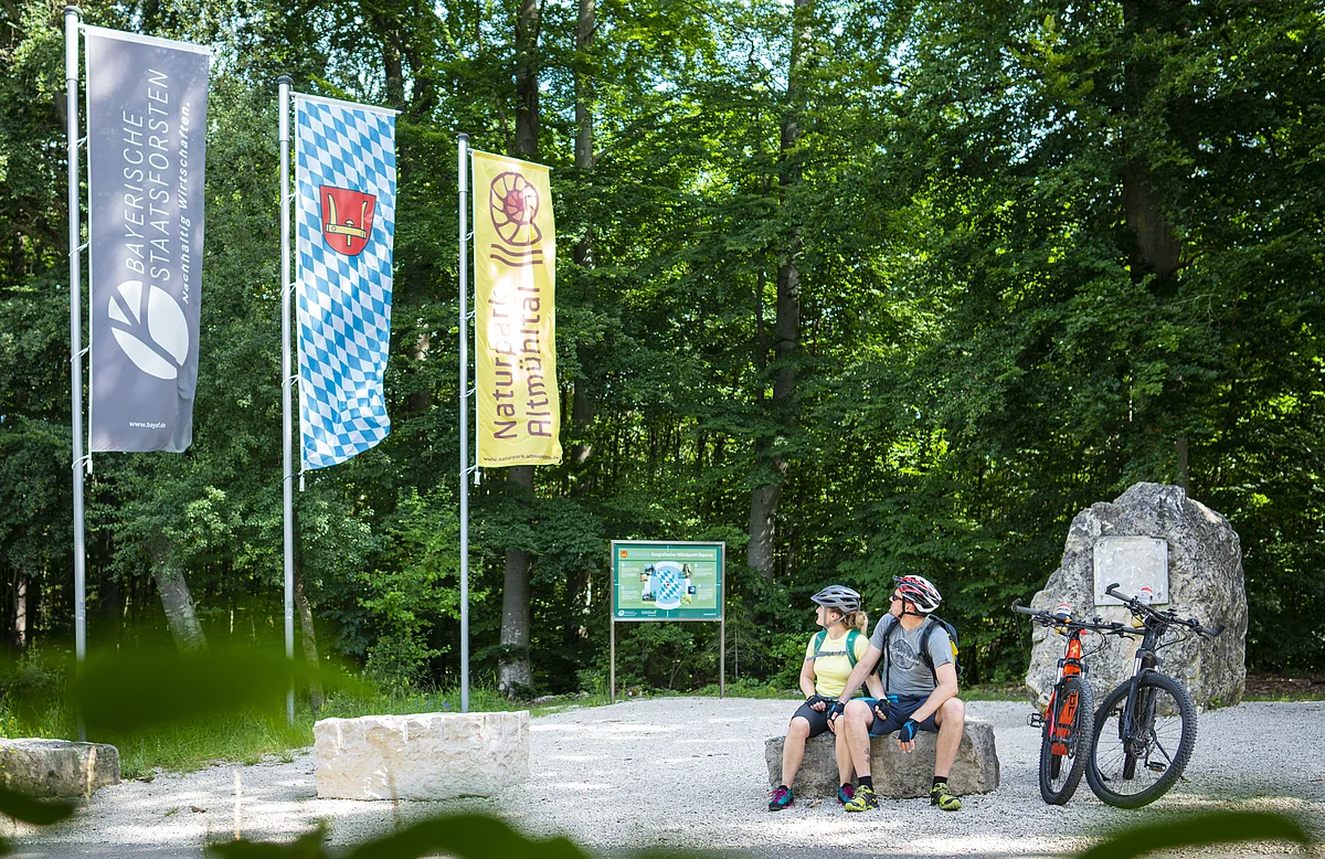Geographischer Mittelpunkt Bayerns Zwei Radfahrer mit Helm sitzen auf Steinbank im Wald neben zwei Mountainbikes und drei Fahnen.